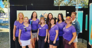 Eight Art Harvest Committee Members posing with Michelle Mardis' in front of her fine art work.